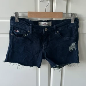 Mörkblå jeansshorts från Hollister - Snygga mörkblå lågmidjade jeansshorts från Hollister med råa, fransiga kanter och slitningar på ena benet. Klassisk femficksmodell med broderad logga på fickan och läderpatch bak 💙 Kan fixa fler bilder!