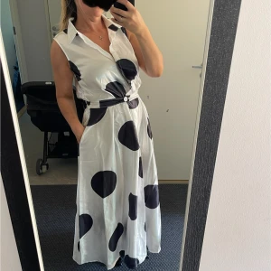 Vit långklänning med stora svarta prickar - Snygg ärmlös långklänning i vit med stora svarta prickar. Klänningen har krage, knappar framtill och ett matchande bälte i midjan. Materialet är glansigt och ger en lyxig känsla. Perfekt för dig som vill sticka ut med ett coolt mönster.Tillgängliga storlekar 38-40 och 42-44 Helt ny.Skriv vilken storlek vill du ha.På mig är 38-40 på bilderna