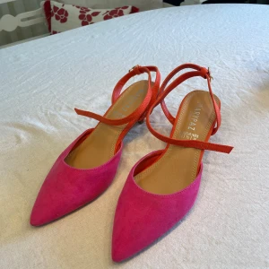 Marypaz pumps i rosa och orange mocka - Färgstarka pumps från Marypaz i mocka med spetsig tå och låg klack. Skorna har en snygg kombination av rosa framtill och orange baktill samt smala remmar med spänne runt ankeln. Perfekta för dig som vill sticka ut med din stil.
