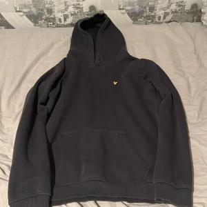 Svart hoodie från Lyle & Scott - Säljer en svart hoodie från Lyle & Scott med klassisk gul logga på bröstet. Tröjan har huva, känguruficka och ribbade muddar. Perfekt för dig som gillar en enkel och stilren look. Materialet är mjukt och skönt, troligen bomullsmix.