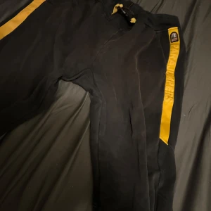Svarta fleecebyxor Parajumpers XXL - Svarta fleecebyxor från Parajumpers i storlek XXL. Byxorna har en gul sidoremsa med Parajumpers-logga och coolt spänne i midjan. Perfekta för chill eller när du vill ha något riktigt bekvämt och snyggt på dig. Färgen är urtvättat! 