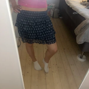 Mönstrad blå kortkjol från Lindex - Supersöt blå kortkjol från Lindex med vitt mönster och volanger. Stor i storleken. Kjolen har resår i midjan för skön passform och är perfekt för dig som gillar en luftig och lätt stil. Passar dig som vill ha en chill och snygg look till sommaren.