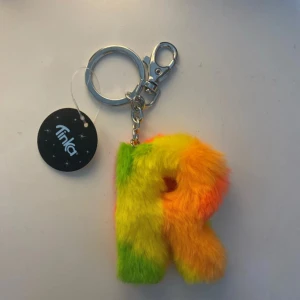 Färgglad fluffig R-nyckelring från Tinka - Superfin och mjuk nyckelring från Tinka i form av bokstaven R. Den är täckt av fluffigt syntetmaterial i neonfärger som gul, grön och orange. Perfekt att hänga på väskan eller nyckelknippan för en färgklick.