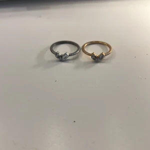 Två ringar i silver och guld med sten - Säljer två snygga ringar, en i silverfärg och en i guldfärg, båda med en glittrande klar sten i mitten. Ringarna har en tunn, räfflad ringband och en elegant, modern design som passar perfekt för att lyfta din stil.