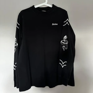 Svart långärmad tröja från Dickies - Svart långärmad tröja från Dickies med vita tryckdetaljer på ärmarna, inklusive en cool orm och grafiska mönster. Tröjan har en klassisk rund hals och är gjord i mjuk bomull, perfekt för en avslappnad streetstyle-look.