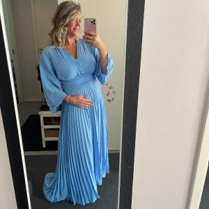 Ljusblå plisserad långklänning - Superfin ljusblå långklänning med plisserad kjol och vida ärmar. Klänningen har v-ringning och markerad midja med bred linning. Perfekt för dig som vill ha en flowy och elegant look. Materialet känns lätt och luftigt.