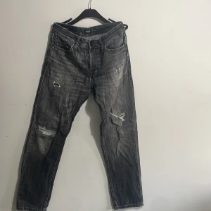Svarta slitna jeans Jack & Jones - Svarta jeans från Jack & Jones i tvättad look med slitningar och trasiga detaljer på benen. Klassisk femficksmodell med knappgylf och smal passform. Perfekta för dig som gillar en edgy och avslappnad stil.