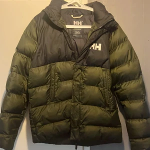 Grön och svart pufferjacka Helly Hansen - Säljer en grön och svart pufferjacka från Helly Hansen med huva och dragkedja. Jackan har stora fickor framtill och HH-logga på bröstet och ryggen. Perfekt för kalla dagar och riktigt snygg streetstil.