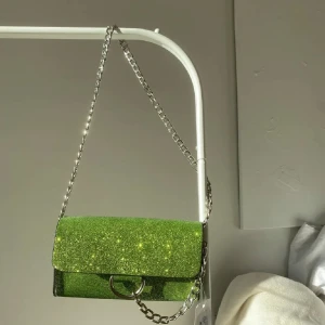 Grön glittrig axelväska från NLY - Säljer en grön glittrig axelväska från Nelly. Väskan har en silvrig kedjerem och ett stilrent spänne framtill. Perfekt för dig som vill sticka ut med en färgstark accessoar. Rymmer mobil, plånbok och småsaker💚