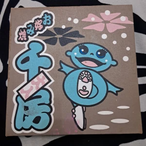 Happy Chappy - En färgglad och lekfull tavla av en blå axolotl med stora ögon och ett glatt uttryck. Perfekt för dig som gillar söt och unik konst med japanska influenser. Passar utmärkt som dekoration i ungdoms- eller studentrummet. Texten är på kinesiska (vet ej vad den säger). Den har några fläckar på baksidan, men syns ej genom