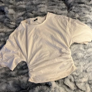 Vit kortärmad topp med rynkdetalj - Säljer en vit topp med korta ärmar och snygg rynkdetalj på båda sidor. T-shirten har en avslappnad passform och är gjord i mjuk bomull, perfekt för en chill och stilren look.