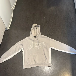 Beige/grå hoodie från Lorenzo med tryck - Snygg beige/grå hoodie från Lorenzo veratti med broderad logga framtill och citat på ryggen. Tröjan har huva med snörning, känguruficka och vita detaljer längs ärmarna. Perfekt för en chill och avslappnad stil.