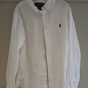 Vit skjorta från Ralph Lauren - Klassisk vit skjorta från Ralph Lauren med broderad logga på bröstet. Skjortan har lång ärm, knappar framtill och en snygg krage. Custom fit-modell i mjuk bomull som passar perfekt till en clean och stilren look.