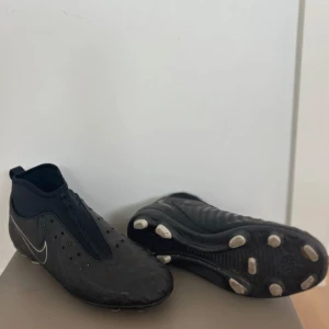 Svarta Nike Phantom GX 2  - Snygga svarta Nike fotbollsskor med vit swoosh och klassisk design. Skorna har en strömlinjeformad form, täckt snörning och dobbar för bra grepp på planen. Ovandelen är i syntetmaterial och skon har en tight passform runt ankeln för extra stöd.