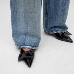 Svarta lackpumps med rosett från NA-KD - Snygga svarta pumps i lack från NA-KD med spetsig tå, låg klack och öppen häl. Skorna har en stor dekorativ rosett framtill som ger en cool och trendig vibe. Skorna är helt oanvända och ligger kvar i den befintliga lådan. De säljdes då jag köpt dem i för stor storlek och har därför ingen användning av dem💞💞