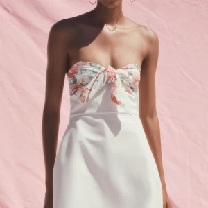 Vit axelbandslös kortklänning med rosett - Superfin axelbandslös kortklänning i vitt med blommigt mönster och stor rosett framtill. Klänningen har en figurnära passform upptill och lätt utsvängd kjol. Perfekt för sommarens alla tillfällen! Säljer då den ej passar mig