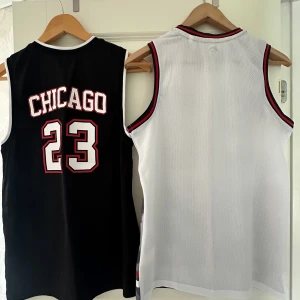 Chicago basketlinnen från Tenth - Två basketlinnen från Tenth, ett svart med CHICAGO och nummer 23 i vitt och rött, och ett vitt med grått camomönster och nummer 90. Båda är ärmlösa, har V-ringning med randiga muddar och är gjorda i luftig polyester mesh. Perfekta för basket eller streetwear i storlek 164