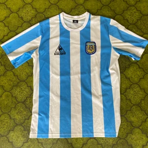 Argentina Maradona tröja  - Klassisk Argentina fotbollströja med breda blå och vita ränder, officiellt AFA-märke och Le Coq Sportif-logga på bröstet. På ryggen står det Maradona och nummer 10. Tillverkad i polyester, kortärmad och med rund hals. Perfekt för dig som älskar retro och fotbollshistoria. Size L 