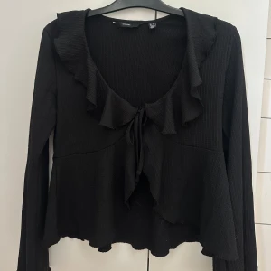 Svart volangblus med knyt från H&M - Supersöt svart blus från Vero Moda med volangkant längs den djupa v-ringningen och knyt framtill. Endast använd ett fåtal gånger. Hör av er vid frågor om pris eller annat💖