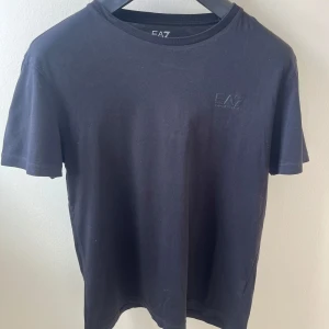 Svart t-shirt från EA7 Emporio Armani - Svart t-shirt från EA7 Emporio Armani med diskret logga på bröstet. Klassisk rund halsringning och korta ärmar. Tillverkad i mjuk bomull för en skön känsla och enkel stil. Perfekt för dig som gillar stilrena och snygga basplagg.