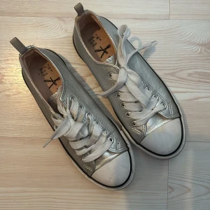 Silvriga sneakers - Snygga sneakers i metallic silver med vit tå och vita snören. Klassisk låg modell med svarta ränder på sulan och rund tå. Ovandelen är i syntetmaterial som ger en cool glansig look. Perfekta för dig som vill sticka ut lite extra i vardagen.