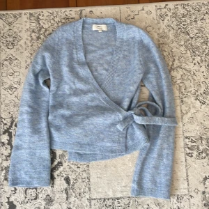 Ljusblå omlottkofta från Gina Tricot - Supermysig ljusblå omlottkofta från Gina Tricot i ullblandning. Koftan har långa ärmar, ribbade muddar och knytband i sidan för en snygg passform. Perfekt att slänga på sig när det är lite kyligt ute.