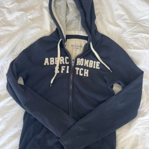 Marinblå hoodie Abercrombie & Fitch - Mysig marinblå hoodie från Abercrombie & Fitch med vit broderad logga framtill. Tröjan har huva med vita snören, dragkedja och kängurufickor. Perfekt för chill dagar och snygg till jeans eller mjukisbyxor.
