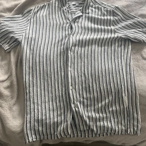 Randig skjorta från SMOG, relaxed fit - Kortärmad skjorta från SMOG i relaxed fit, med svartvita vertikala ränder och knäppning framtill. Skjortan är tillverkad i ett luftigt tyg som passar perfekt för varmare dagar. Klassisk krage och stilren design som ger en chill vibe.