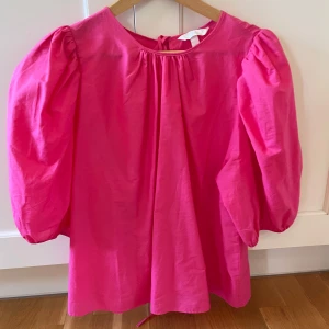 Rosa blus med puffärm från H&M - Superfin rosa blus från H&M med puffiga trekvartsärmar och snygga knytband i ryggen. Blusen har en lös passform och är gjord i mjuk bomull, perfekt för dig som gillar färg och volym i din outfit.