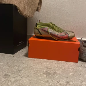 Nike fotbollsskor i grönt och vitt - Säljer ett par Nike fotbollsskor med grön ovandel, vita och röda detaljer samt svart Nike-logga. Skorna har snörning och är designade för spel på gräs med dobbar undertill. Perfekta för dig som vill sticka ut på planen.