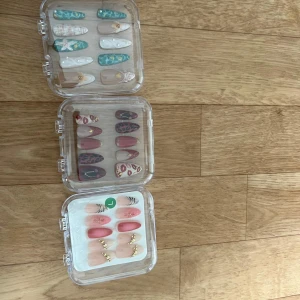 Färgglada lösnaglar i tre askar - Tre askar med lösnaglar i olika designer: turkos och vit med snäckskal och pärlor, vinröd och nude med leopardmönster och gulddekor, samt rosa och nude med blommor och gulddetaljer. Naglarna är långa och har både spetsig och rundad form.