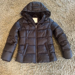 Svart pufferjacka från Tommy Hilfiger - Svart pufferjacka från Tommy Hilfiger med huva och dragkedja framtill. Den är varm och skön perfekt nu när det börjar närma sig höst och vinter. Säljer den då den är för liten och inte används.
