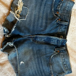 Levi's blå jeansshorts med slitningar - Snygga blå jeansshorts från Levi's med råa, fransiga kanter och slitningar framtill. Klassisk femficksmodell med Levi's-logga på bakfickan. Perfekta för sommaren och ger en avslappnad vibe till din outfit.