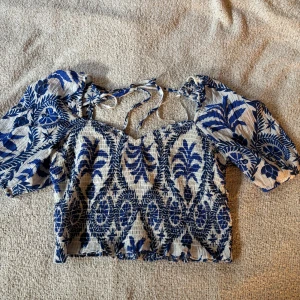 Blommig croppad blus från H&M - Superfin croppad blus från H&M med blått och vitt blommönster. Toppen har puffiga korta ärmar, smockad struktur och söt knytning i nacken💞Blusen är endast använd en gång och därför i nästan nytt skick!