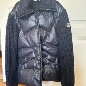 Marinblå Moncler pufferjacka med stickade ärmar - Säljer en marinblå pufferjacka från Moncler med stickade ärmar och hög krage. Jackan har dragkedja framtill, två fickor och Moncler-logga på ena ärmen. Kombinerar glansigt material på kroppen med grovstickade detaljer på ärmar och krage för en riktigt snygg look. Använd fåtal gånger. Inga defekter. Fler bilder kan skickas på begäran, pris går att diskutera. Orginalkartong och alla kvitton finns kvar. Orginalpris 12.000💕