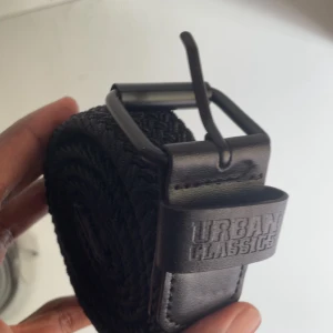 Urban Classics flätade bälten svart & grå - Två snygga flätade bälten från Urban Classics, ett i svart och ett i grått. Båda har stilrena metallspännen och detaljer i konstläder med präglad logga. Perfekta för att lyfta din outfit med en clean och modern vibe.