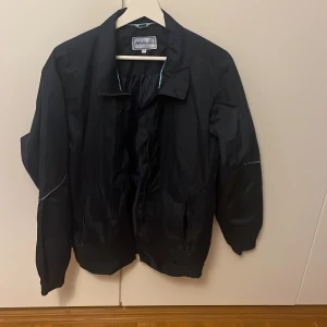 Mörkblå vindjacka från Börjes Tingsryd - Mörkblå vindjacka från Börjes Tingsryd i storlek M. Jackan har dragkedja framtill, två fickor med dragkedja och diskreta reflexdetaljer på ärmarna. Klassisk krage och elastiska muddar vid ärmslut och nederkant. Perfekt för blåsiga dagar.