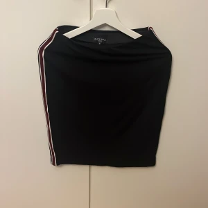 Svart kortkjol Brave Soul med ränder - Svart kortkjol från Brave Soul med sportiga vita och vinröda ränder längs sidorna. Kjolen har en tight passform och är perfekt för dig som gillar en enkel men snygg stil. Materialet känns mjukt och stretchigt.