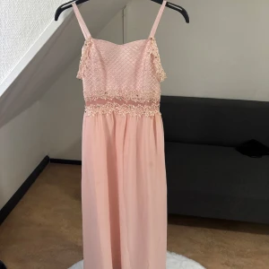 Ljusrosa långklänning med spetsdetaljer - Superfin ljusrosa långklänning med smala axelband och vackra spetsdetaljer upptill. Klänningen har ett flowy fall, markerad midja och dragkedja i ryggen. Perfekt för dig som vill ha en romantisk och elegant look.