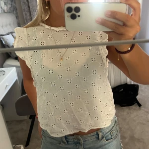 Vit broderad blus med volangärm - Superfin vit blus med broderade hålmönster och små volanger vid ärmslut. Toppen är ärmlös med en lätt croppad passform och rund halsringning. Perfekt till jeans eller kjol för en fräsch och somrig look.