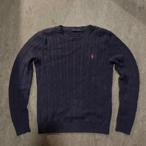 Mörkblå kabelstickad tröja Ralph Lauren - Kabelstickad mörkblå tröja från Ralph Lauren med klassisk rund halsringning och rosa broderad logga på bröstet. Tröjan är storlek Medium (M) och är i väldigt bra skick. 
