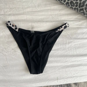Svarta bikiniunderdelar med leoparddetalj - Snygga svarta bikiniunderdelar med högt skuren modell och leopardmönstrade band i sidorna. Perfekt för stranden eller poolen, ger en trendig och cool vibe. Materialet känns mjukt och följsamt mot huden.