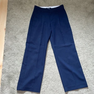 Mörkblå randiga kostymbyxor Gina Tricot - Snygga mörkblå kostymbyxor från Gina Tricot med tunna ljusare blå linjer. Byxorna har raka ben, hällor för bälte och klassisk passform. Mycket bra skick, de är knappt använda. Midjan är 37cm rakt över och inerbenslängd är 77 cm.