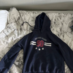Mörkblå hoodie från Gina Tricot XS - Säljer en mörkblå hoodie från Gina Tricot i storlek XS. Tröjan har tryck med texten 'New York League 90' och vita ränder samt stjärnor på bröstet. Klassisk huva med snörning och långa ärmar. Perfekt för en chill och sportig look.