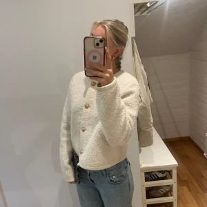 Vit teddykofta  med guldknappar - Supermysig vit teddyjacka med rund hals och snygga guldiga knappar framtill. Jackan är croppad och har lång ärm, perfekt för lager-på-lager. Den fluffiga strukturen ger en trendig vibe och passar till allt från jeans till kjol.