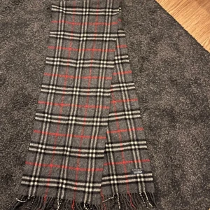 Rutig grå halsduk från Burberry - Snygg halsduk från Burberry i klassiskt rutigt mönster med grå bas, svarta, vita och röda ränder. Gjord i 100% lammull och har fransar i ändarna. Perfekt accessoar för att lyfta din outfit med en tidlös vibe.