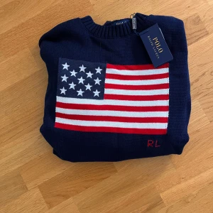 Mörkblå stickad tröja Polo Ralph Lauren - Mörkblå stickad tröja från Polo Ralph Lauren med amerikansk flagga framtill och RL-broderi. Tröjan har rund halsringning, ribbade muddar och är långärmad. Perfekt för dig som gillar klassisk och sportig stil. Obs inte orginal