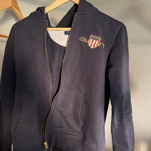 Svart hoodie från Gant med dragkedja - Svart hoodie från Gant med broderad logga och emblem på bröstet. Tröjan har huva, fickor framtill och hel dragkedja. Perfekt för en chill och sportig look. Klassisk passform och mjukt bomullsmaterial.