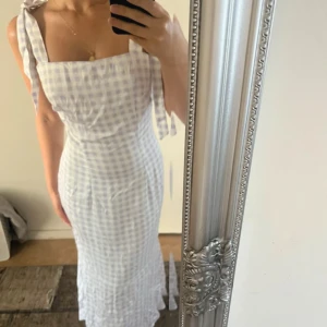 Ljuslila rutig långklänning med knyt - Supersöt långklänning i ljuslila och vitt rutigt mönster. Klänningen har breda axelband med knyt, fyrkantig urringning och figurnära passform som släpper ut i en lätt volang nertill. Perfekt för sommarens alla tillfällen! 