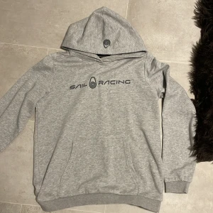 Grå hoodie från Sail Racing - Snygg ljusgrå hoodie från Sail Racing med svart logga på bröstet och en mindre logga på huvan.                                           Hör av dig vid frågor eller mer bilder!!!